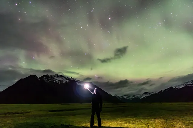 Aurora boreal verde extendiéndose entre nubes sobre montañas oscuras de Islandia, con un cielo lleno de estrellas y una persona en primer plano sosteniendo una luz.